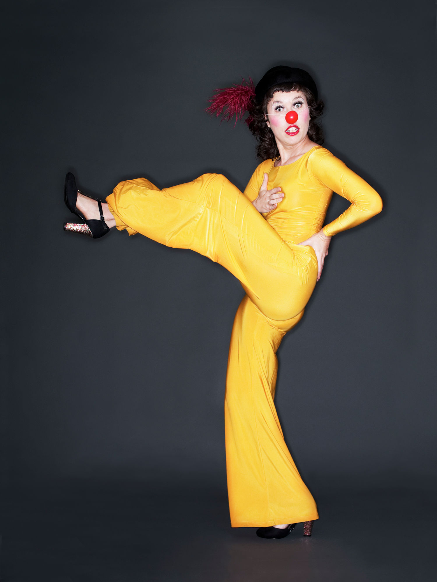 Woman with a red clown's nose in a bright yellow jumpsuit, one leg kicking out in front.