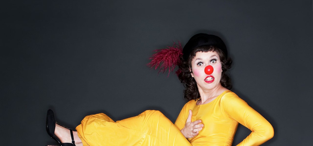 Woman with a red clown's nose in a bright yellow jumpsuit, one leg kicking out in front.