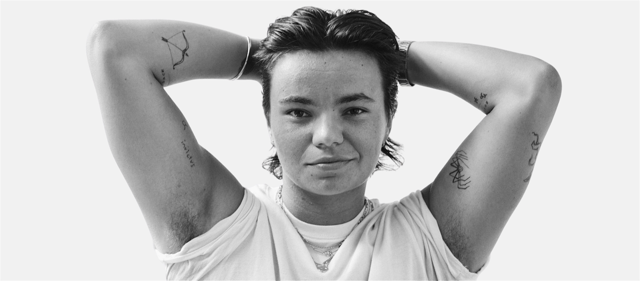 Zoe Terakes shot in black and white facing the camera with their arms up above their head, wearing a white t shirt with sleeves rolled up.