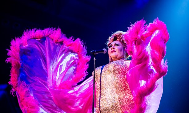 A rather buxsome female impersonator (Trevor Ashley) in gold dress holding large, feathery flag banners, standing before a microphone