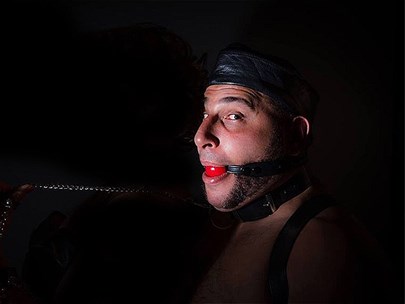 Man bound in leather with a red ball held in their mouth; very dark background