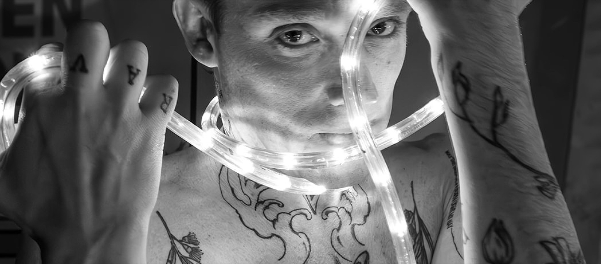 Black and white headshot of non binary performer with heavy tattoos, with arms held up to face a long white rope, light wrapping around hands & neck