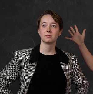 Headshot of a person in a checked jacket, with someone holding one open hand up behind them