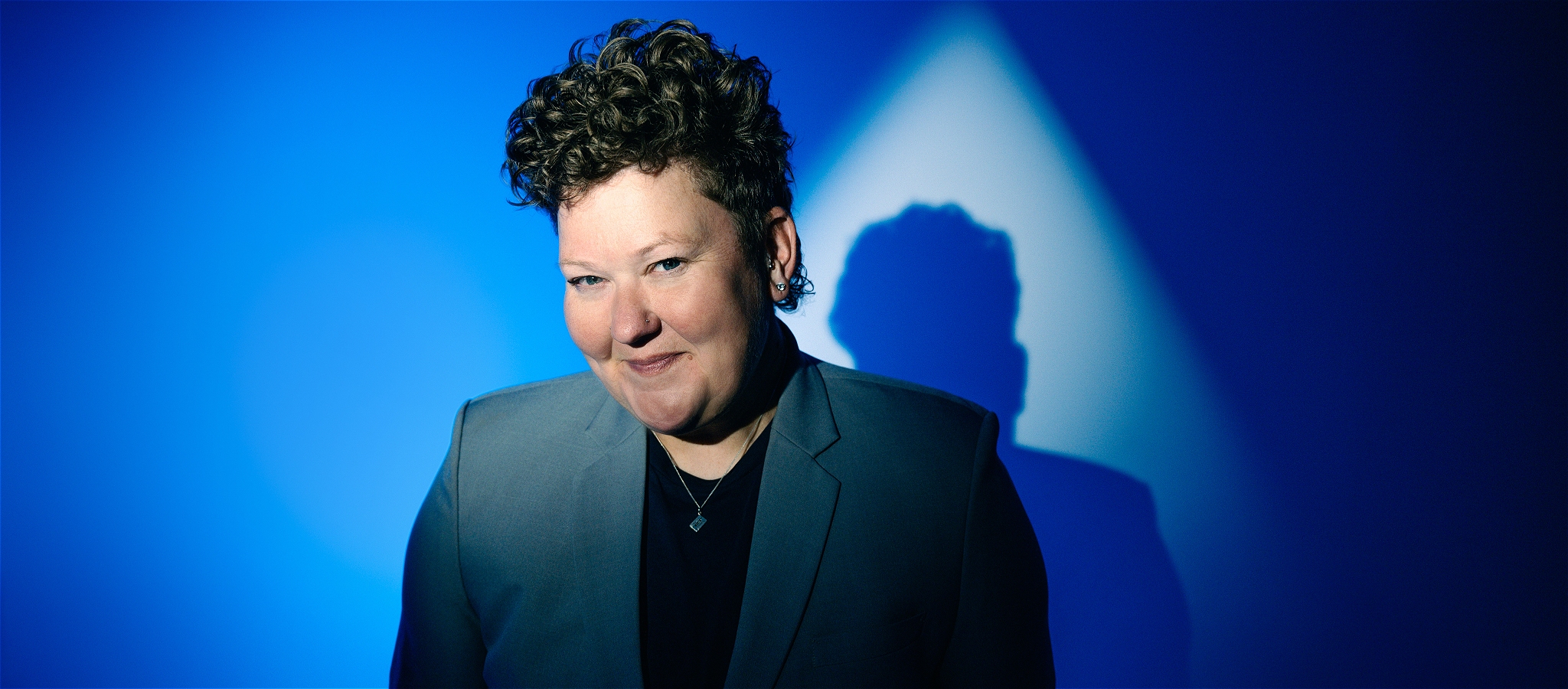 Comedian Kirsty Webeck on Blue Background, with a cheeky grin and curly hair