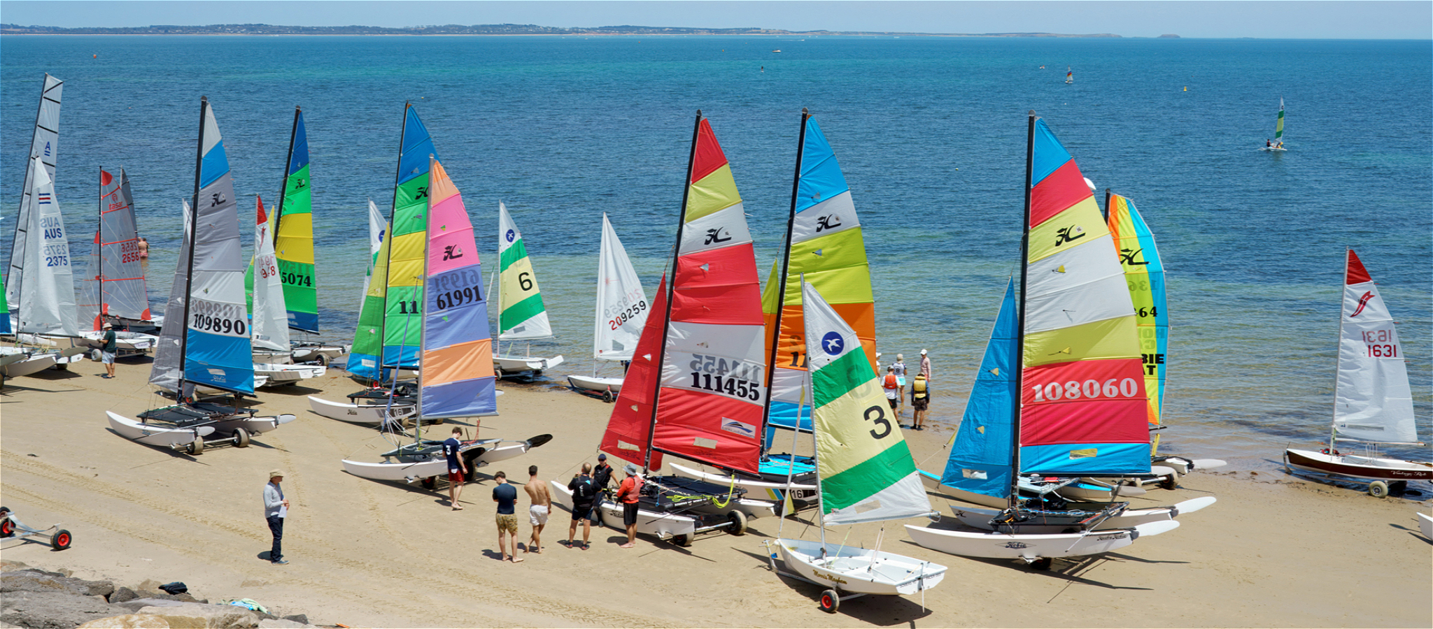 Arouond a dozen boats with colourful sails sit on sand with blue water in the background