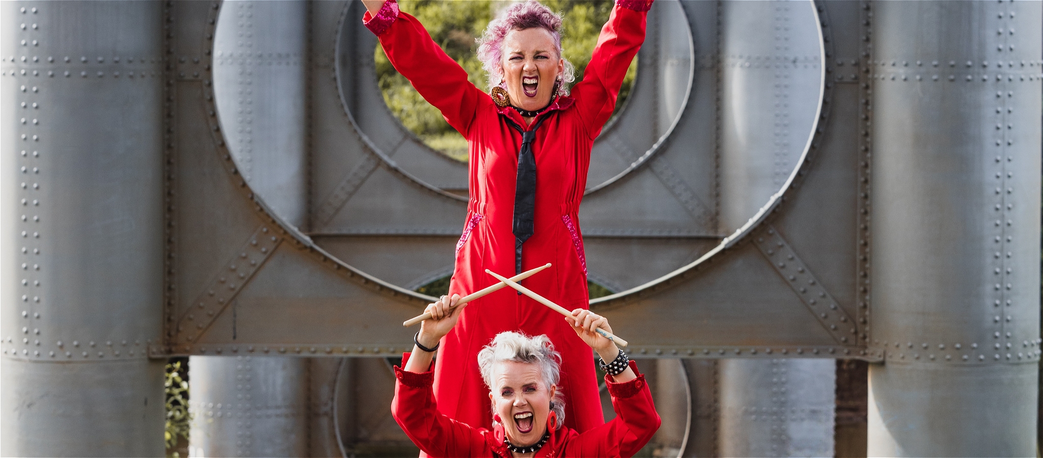 Two women in hot pink short overalls posing with drumsticks crossed and guitar held overhead (off screen)