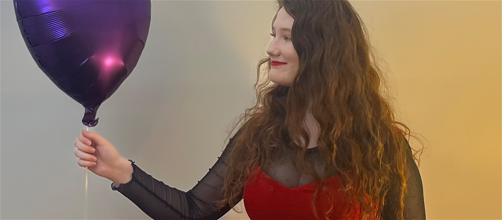 A young woman in a red dress holding and looking at a purple love heart balloon.
