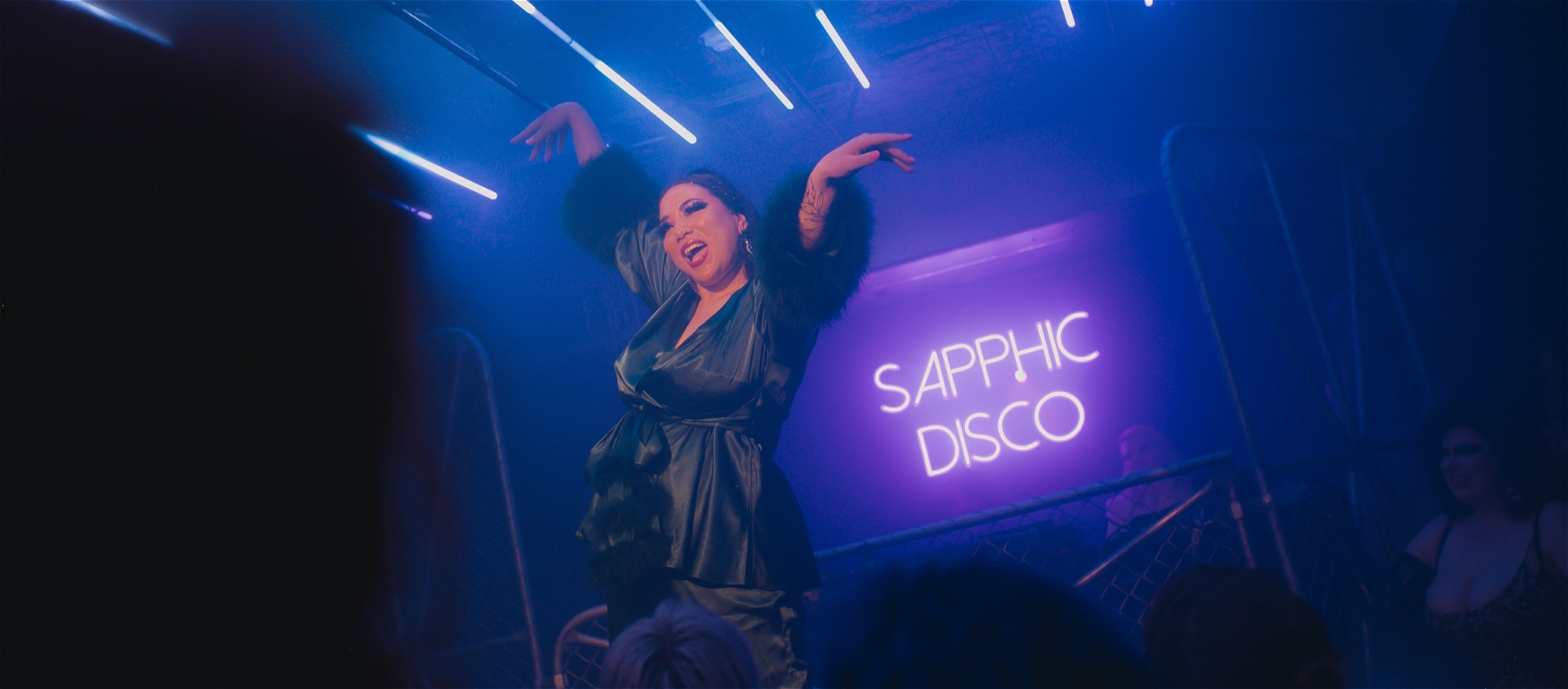 A woman performs in a green silk and fur outfit in front of a neon sign that reads Sapphic Disco
