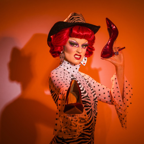 Red-tinged photo of Ruby Slippers sneering while holding stilettos in a menacing manner