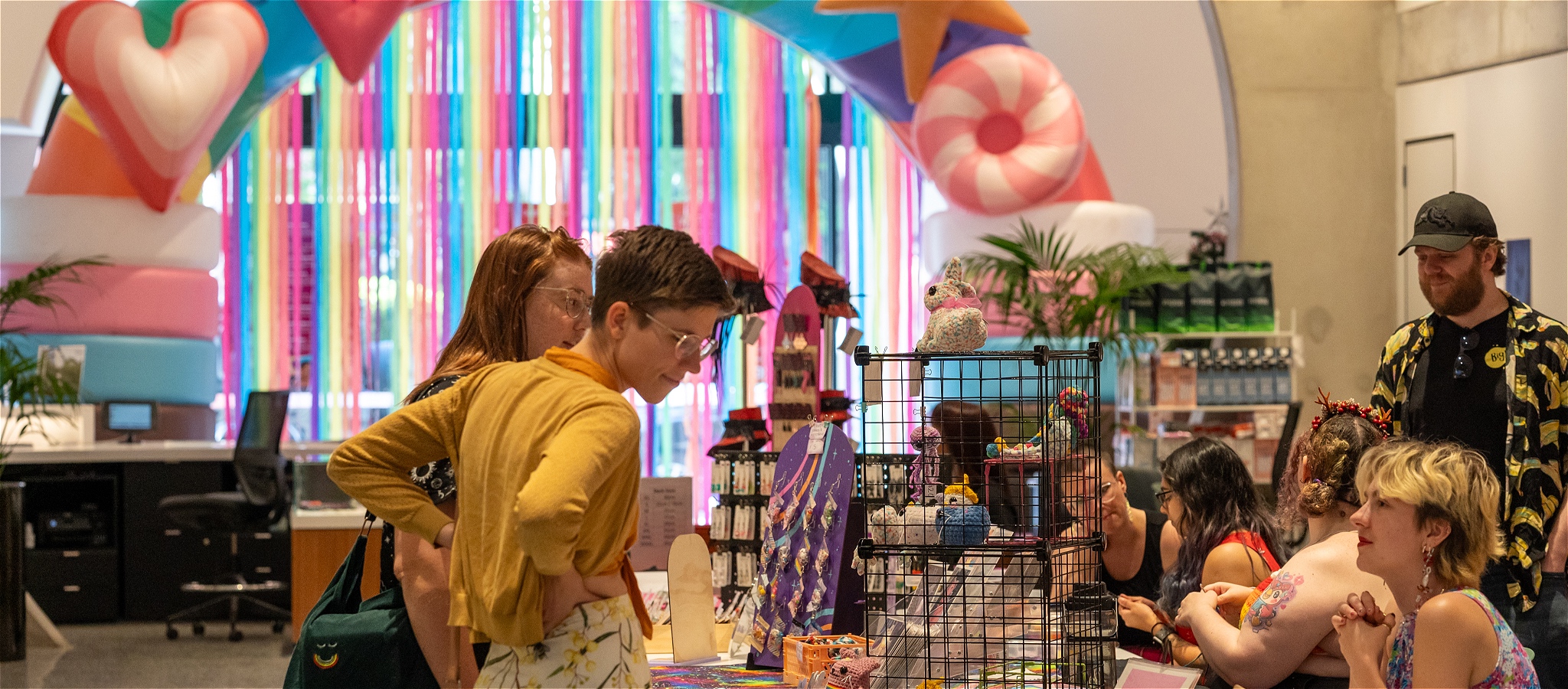 Market-goers browse and chat to queer makers over a stall table with inflatable, rainbow and transgender flag arch and streamers in the background.