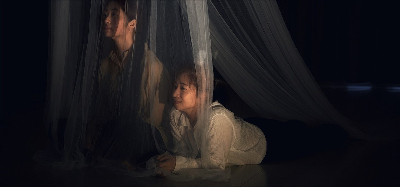 A person lays on their stomach in a dark space, the person next to them sits with crossed legs. They both look beyond white tule that drapes over them