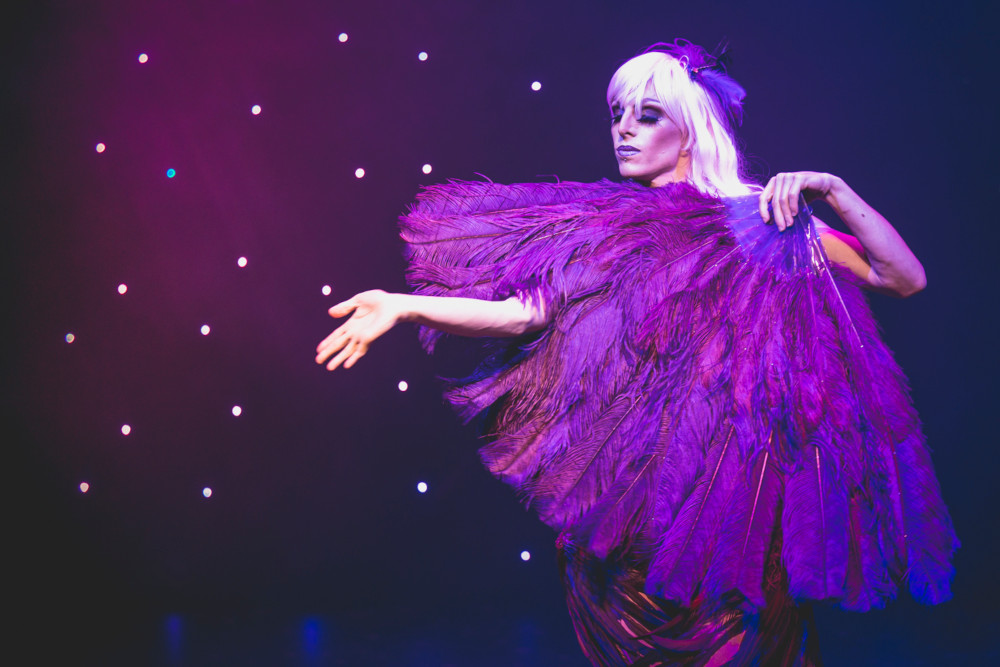 A performer in a "feather-clad fairy" outfit, one hand reaching out; mauve lighting with stars behind
