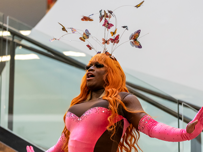 Person of colour in orange wig, and pink and brown decorative tights, with arms stretched out sideways