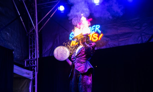 A person creating fire, standing in front of a neon sign that says "Hot Summer Nights"