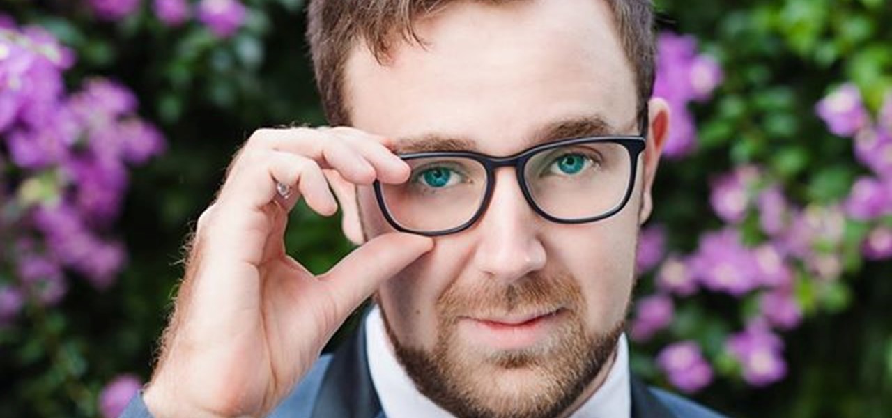 Portrait: Patrick Hayes in grey suit, white shirt, grey blue bow tie, one hand touching his glasses