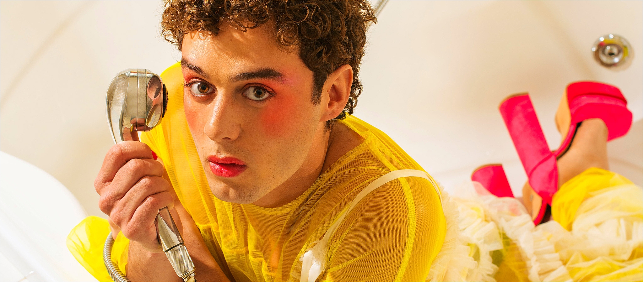 A person in a yellow mesh outfit with pink high heels on lies on their stomach with their head up facing the camera holding a shower head.