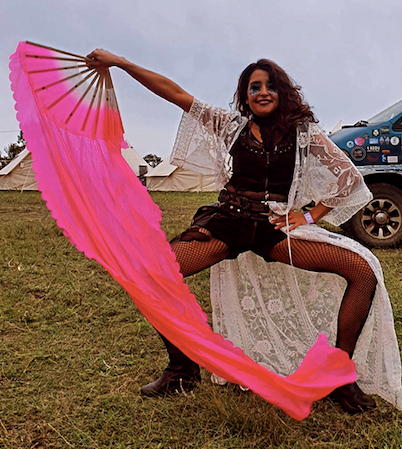 Vivien is waving a pink silk fan and smiling, wearing a white lace jacket and glitter make-up on their face. 