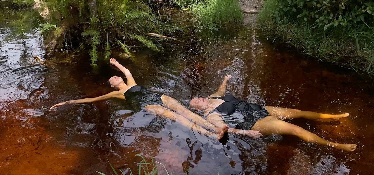 Two people floating face up in a river, wearing black swimsuits with their eyes closed.