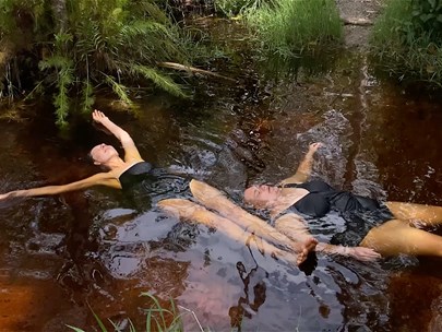 Two people floating face up in a river, wearing black swimsuits with their eyes closed.