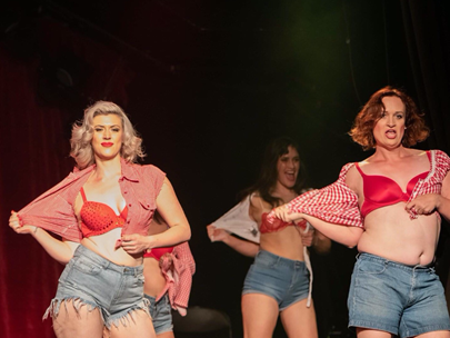 Youngish female-identifying dancers on stage wearing brief shorts and red bras - removing their blouses