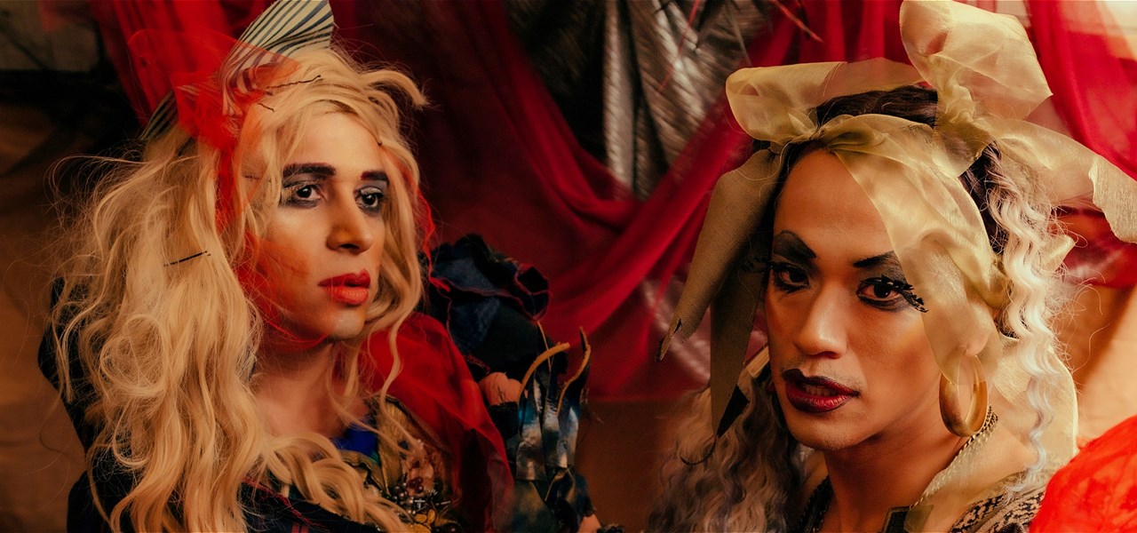A close up of two drag artists. Both of them have messy hair tied up with tulle ribbons, dark eye makeup and red lipstick.