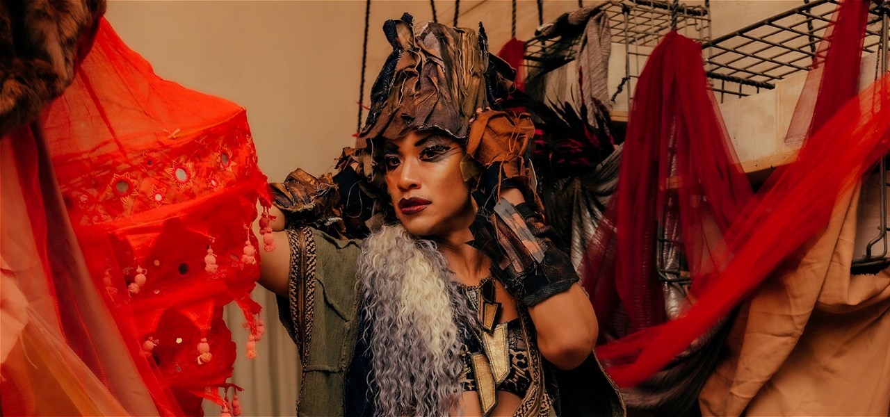 A drag artist posing in front of wire storage and red fabrics. They are wearing a headpiece made of dark mixed fabric that matches their outfit