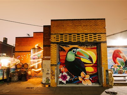 Moon Dog OG's warehouse, adorned with colourful murals at dusk. Fairy lights, a caravan, tables and barrels set the scene.