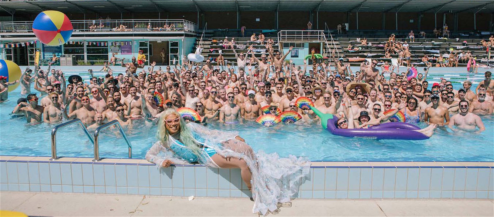 A group photo of a crowd of people celebrating in the pool with a drag queen lounging in the foreground.