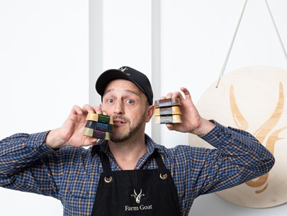 Male-identifying person in Farm Goat apron, holding up 3 bars of soap in each hand