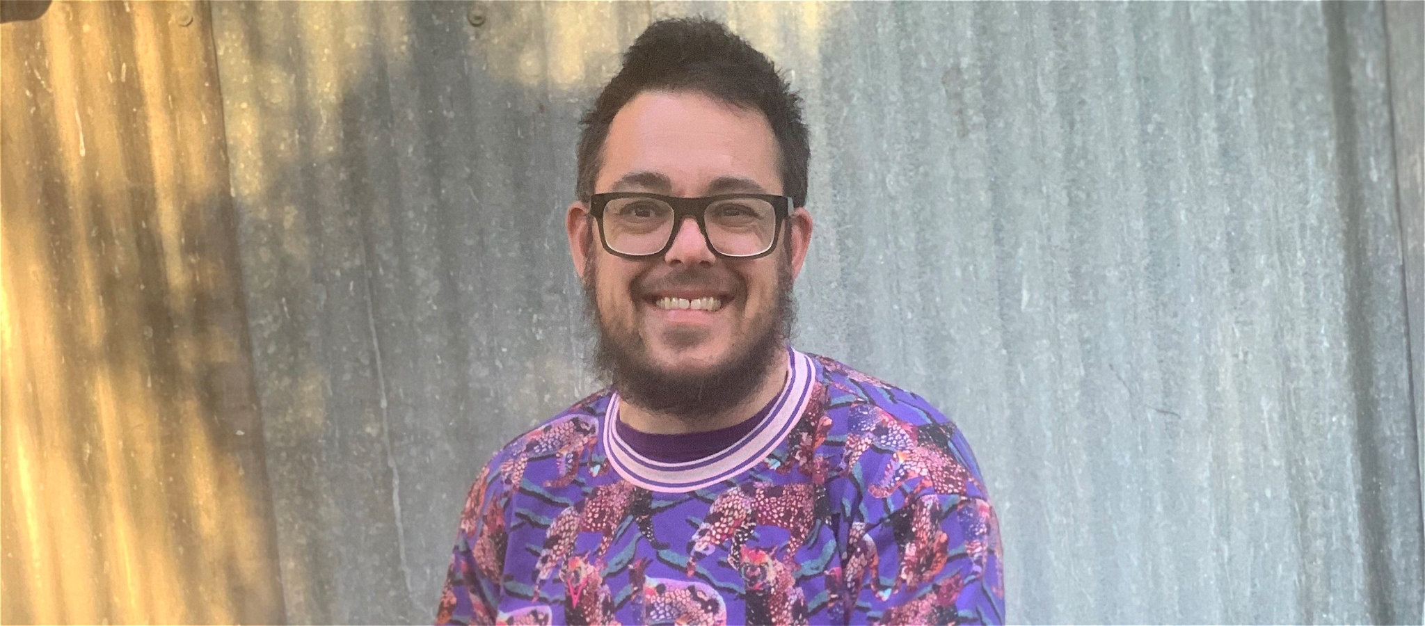 A person with brown hair and beard, wearing glasses and a purple printed top.