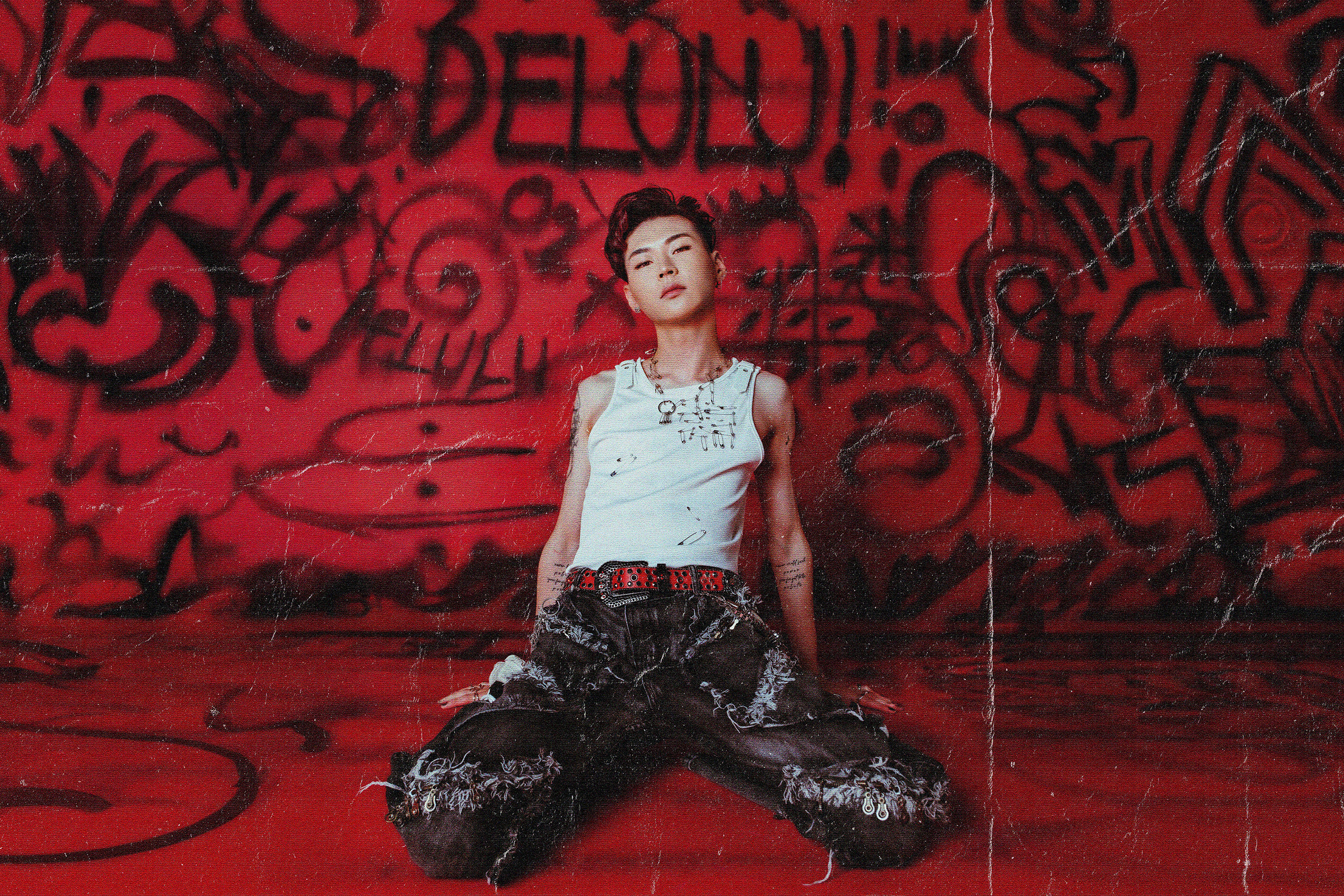 Kevin wearing white singlet and black pants with white designs sitting on the floor in a red lit room with lots of graffiti