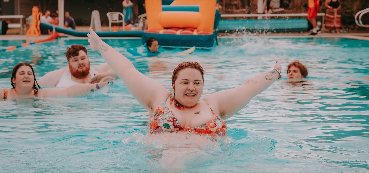 Several young people having fun in the pool at the Pride Pool Party in Sunshine event last year. One of them is leaping out of the water with a smile