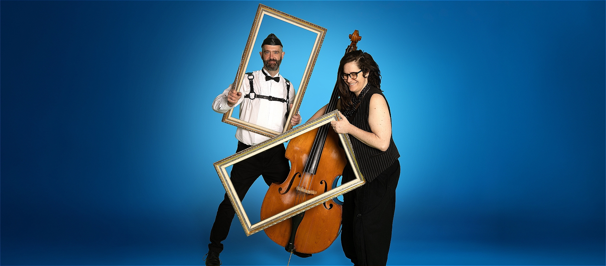 Two people stand behind a gold frame against a blue background holding a double bass