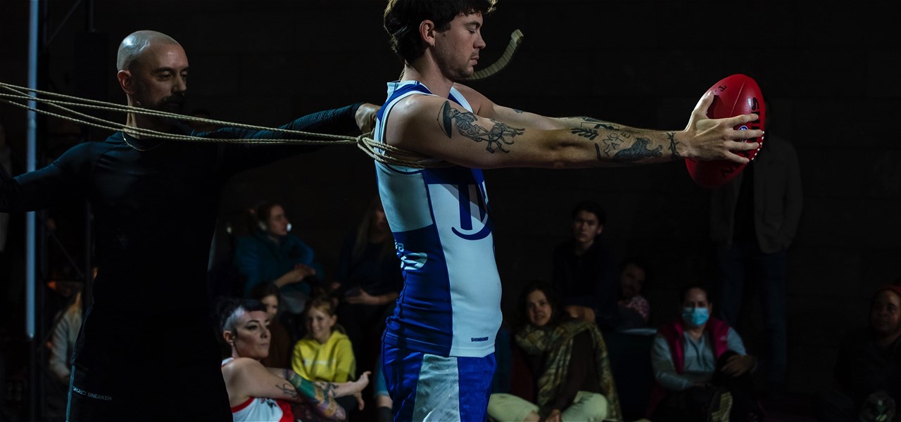 One person with a shaved head and wearing black, is tying another person in rope, who is wearing a white & blue AFL club jersey and holding a football