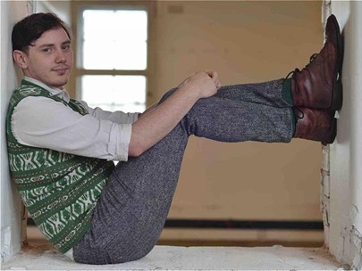 A person in a green patterned knit vest, grey shirt and trousers, is seated in a window like structure with legs in the frame.