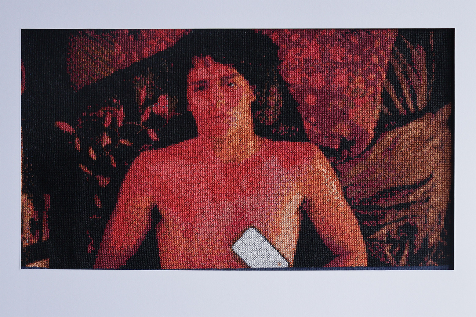 Cross-stitched picture of a bare-chested man lying on some cushions