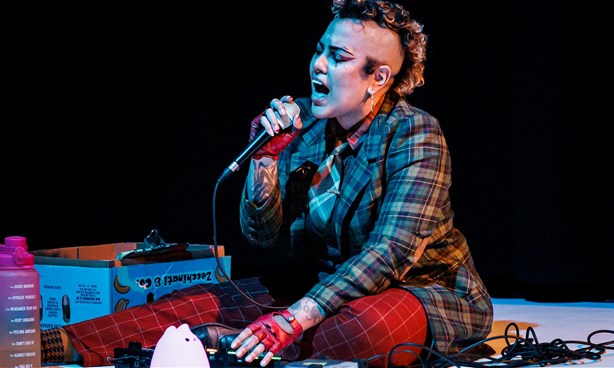 A person with a mohawk sits on the floor singing into a microphone they are holding. They are wearing a brown tartan coat and red pants.