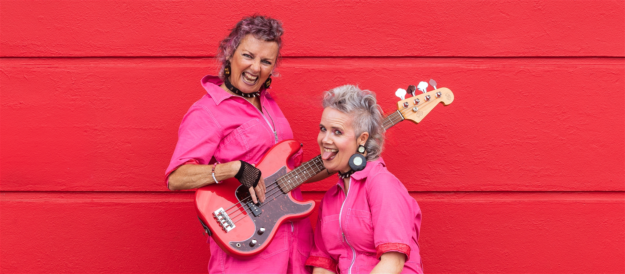 Two women in pink grin at camera, standing in front of a red wall, one holding a red bass guitar, the other trying to lick it.