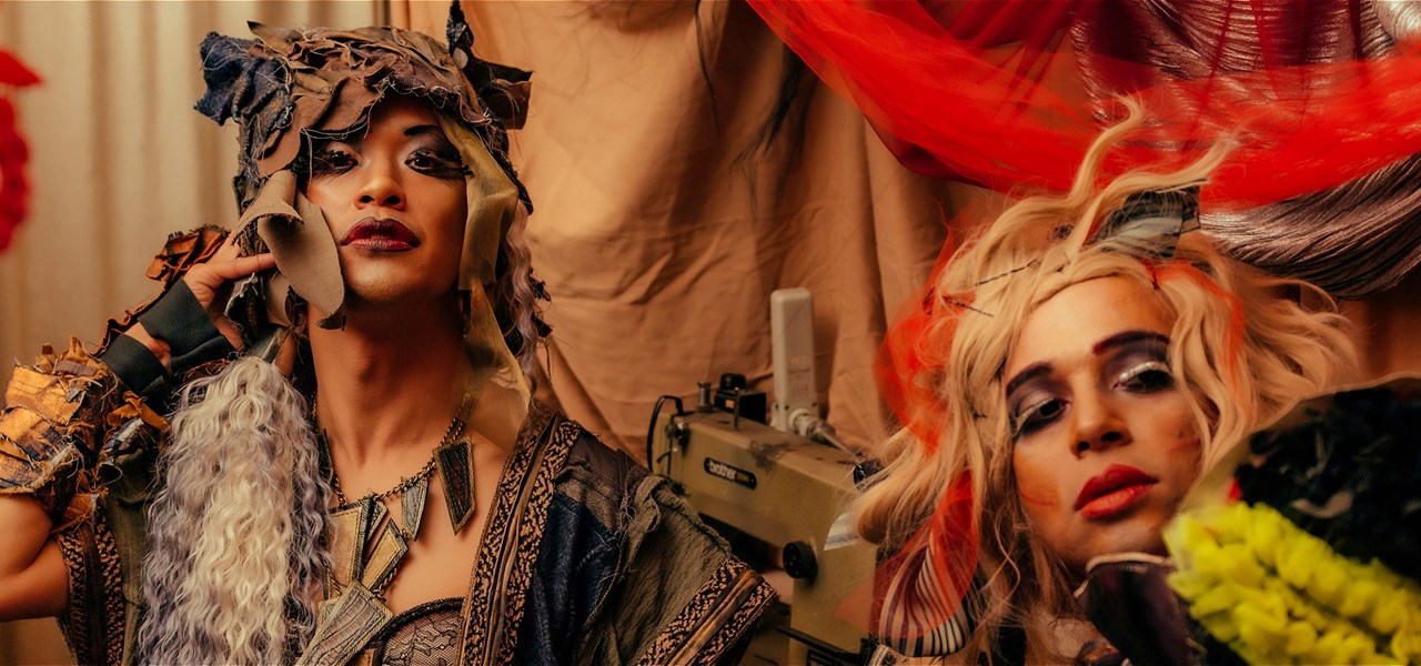 A head and bust portrait of two drag artists posing in garments made of layers of fabric in front of a sewing machine and draped fabric