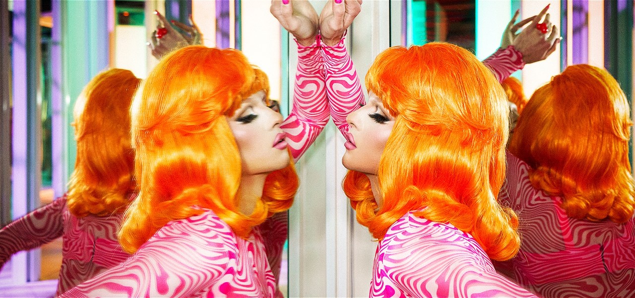 A drag queen in a bright orange 1960s wig stares at herself in the mirror. She is surrounded by mirrors creating six of her