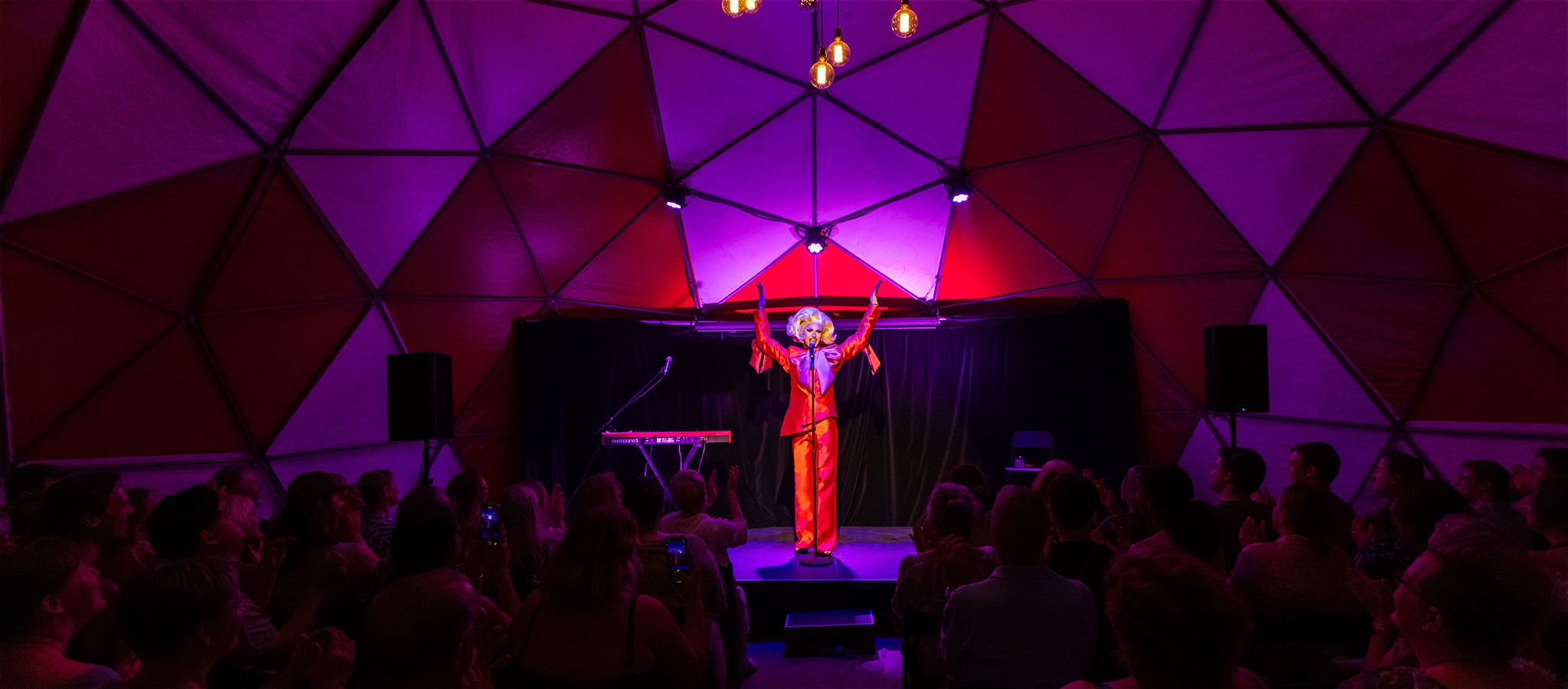 Drag artist stands centre stage in a large cabaret tent with an audience and sings into a mic on a stand, their hands are in the air