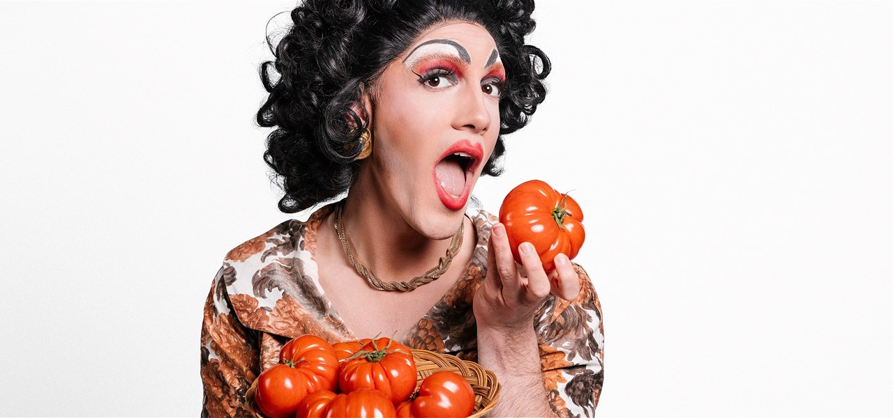 A drag queen, dressed as a Nonna, holds a large red tomato that she is about to take a bite out of.