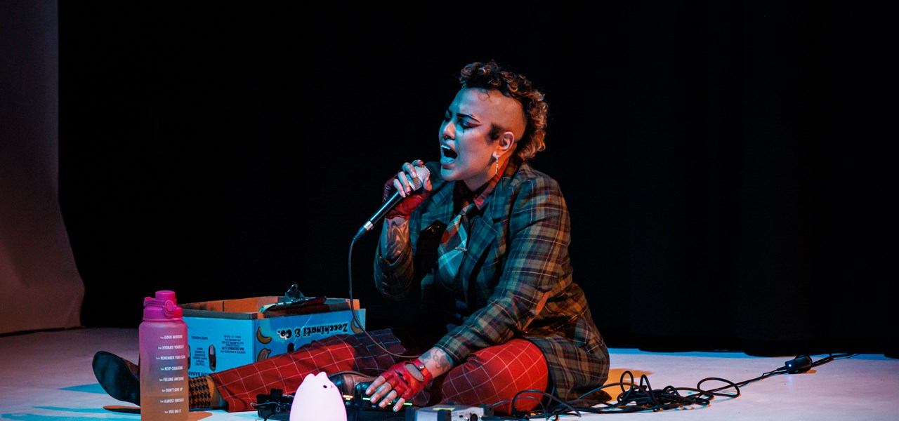 Person dressed in tartan patterned clothes with a "Sioux" haircut, sitting on the floor singing into a mic surrounded by electronic equipment and cables