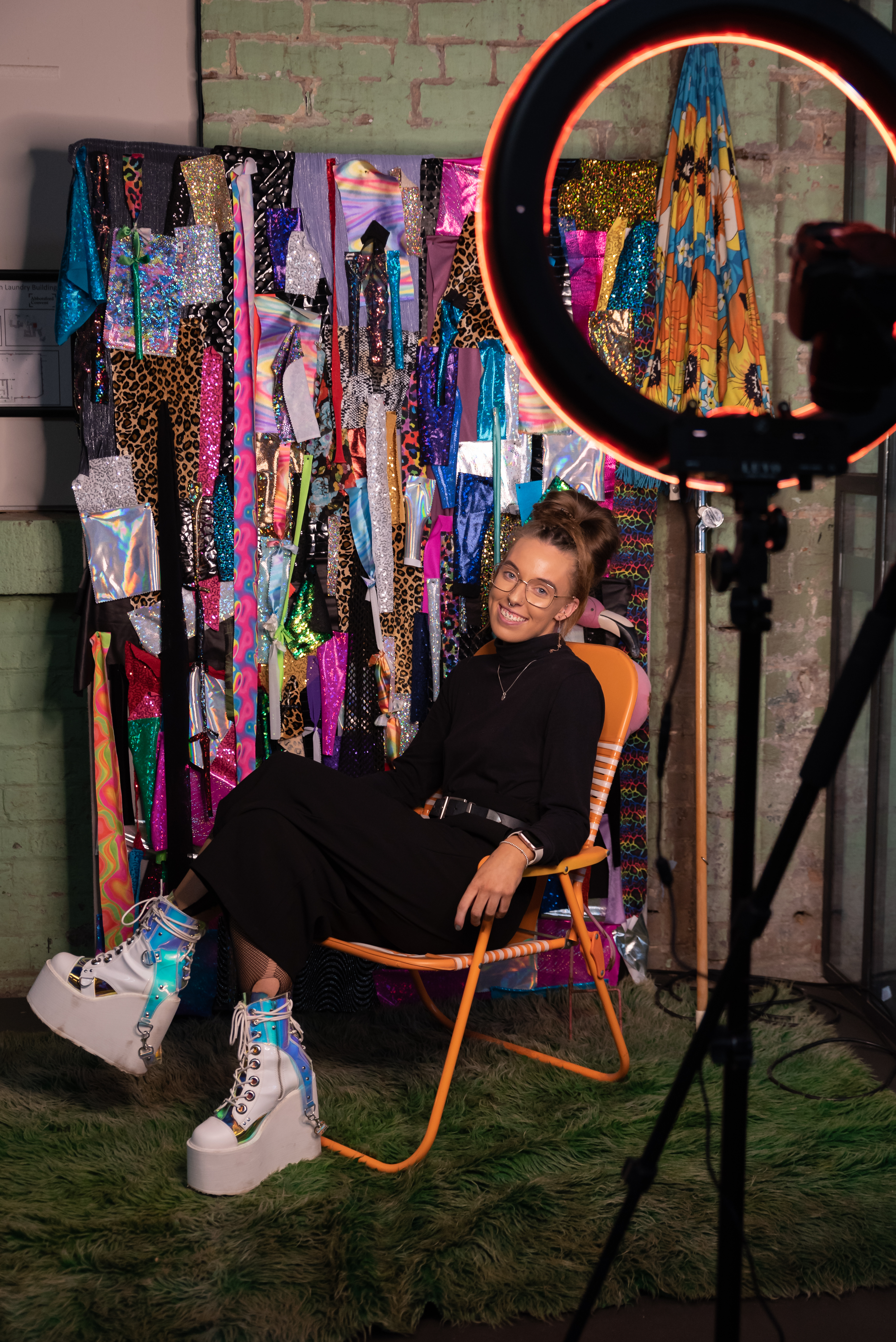 Person dressed in black plus white boots with VERY thick soles sitting in a chair with a rack of colourful clothes behind them
