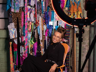 Person dressed in black plus white boots with VERY thick soles sitting in a chair with a rack of colourful clothes behind them