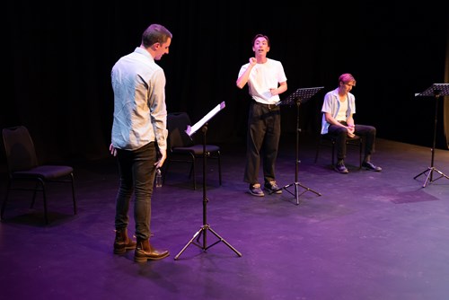 Three people on a stage: one reading from a lectern, one perhaps Auslan interpreting, and one sitting