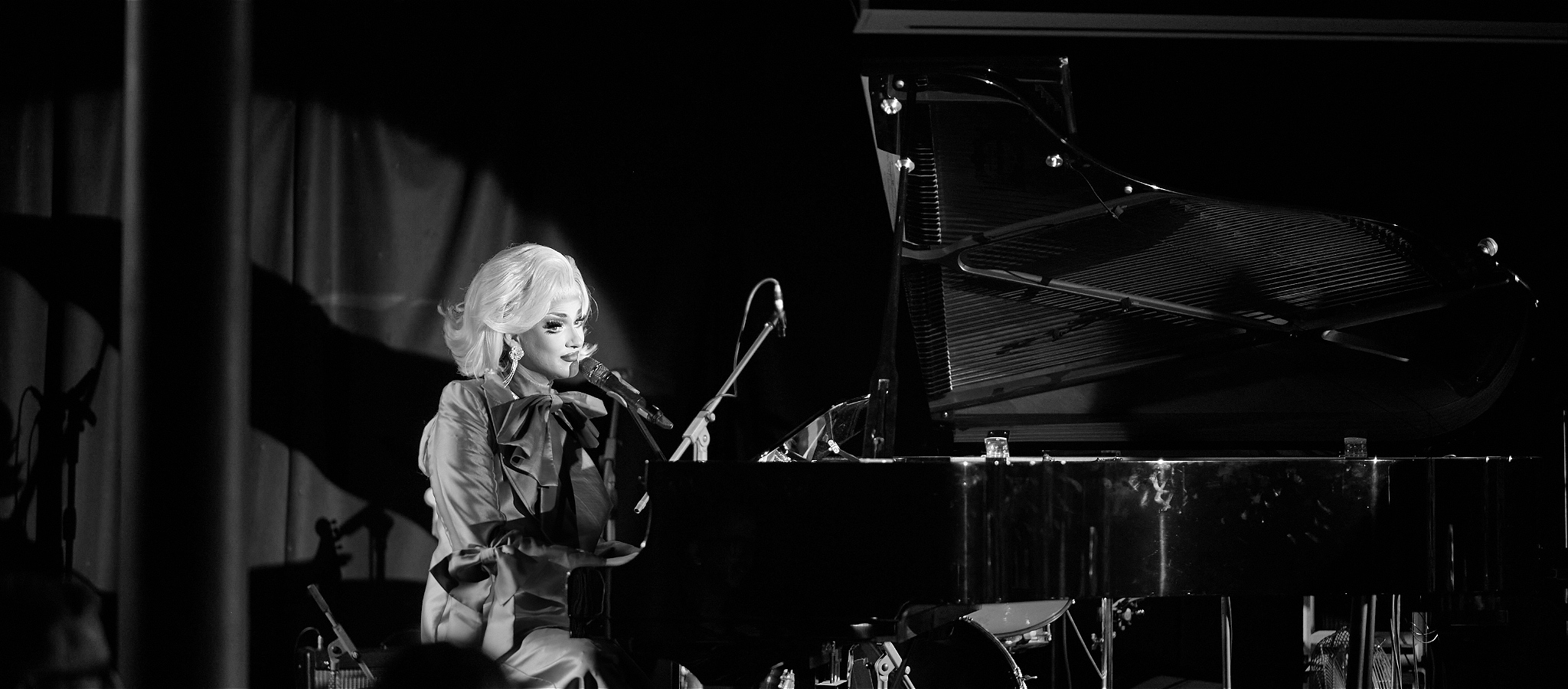 Glamorous drag queen plays grand piano and sings into microphone, she wears a suit and has blonde hair. the image is black and white.