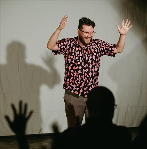 A happy looking Sam on stage, holding up their hands. Silhouettes in the foreground indicate the audience is doing likewise.