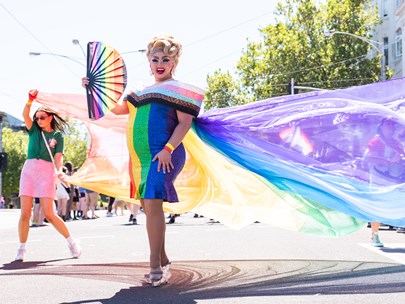 Drag queen holding a rainbow flag with people behind holding a flowing, rainbow coloured sheer, long length of fabric