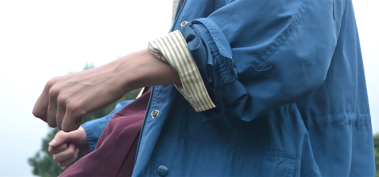 A cropped photograph of a woman wearing a blue trench coat, her arms are bent to hold her clenched fists outwards, as if riding an invisible horse